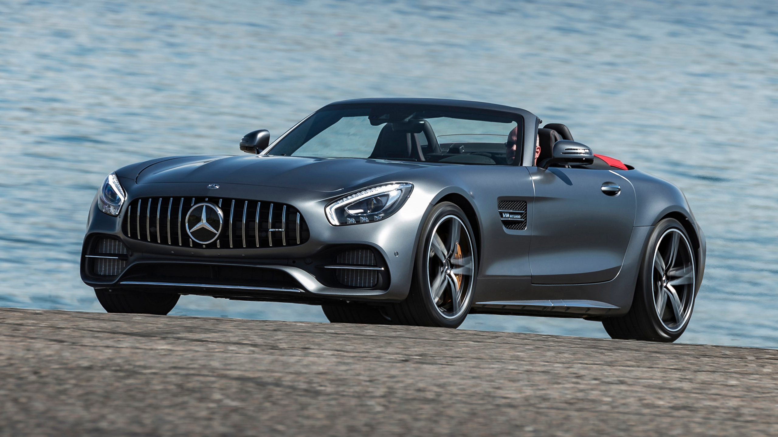Mercedes AMG GT C Roadster gris con capota bajada, en carretera junto al mar.